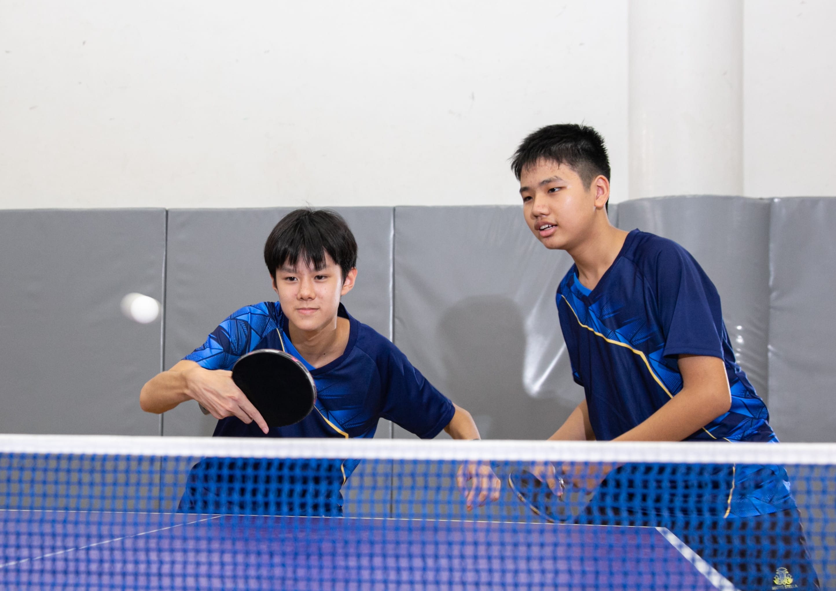 Two men playing table tennis, one in blue swinging a paddle, the other in red watching from the foreground.